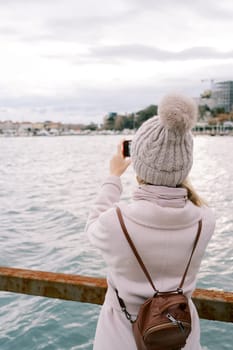Girl with a backpack stands on the pier and takes pictures of the sea coast. Back view. High quality photo