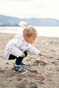 Little girl squats on the beach and picks the sand with a stick. High quality photo