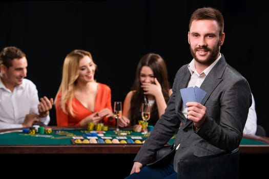 Poker players in casino with cards and chips on black background. A handsome man in the foreground, behind a game table.