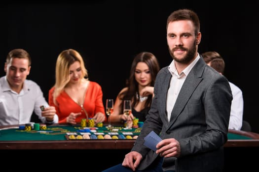 Poker players in casino with cards and chips on black background. A handsome man in the foreground, behind a game table.