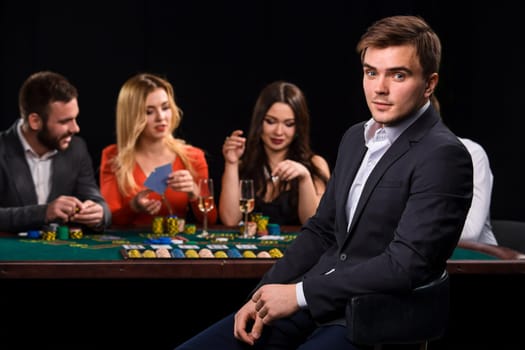 Poker players in casino with cards and chips on black background. A handsome man in the foreground, behind a game table.