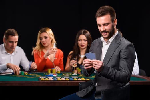 Poker players in casino with cards and chips on black background. A handsome man in the foreground, behind a game table.