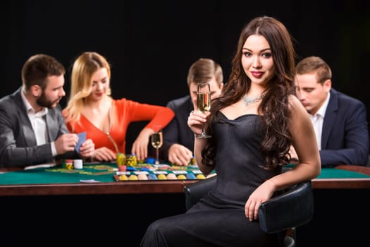 Young people playing poker at the table. Luxury women in dresses with a glass of champagne in hand sitting in the foreground. Casino