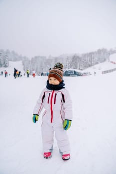 Little smiling girl stands on a snowy ski slope against the backdrop of skiers. High quality photo