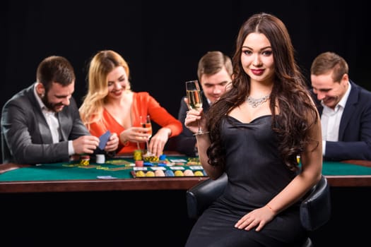 Young people playing poker at the table. Luxury women in dresses with a glass of champagne in hand sitting in the foreground. Casino