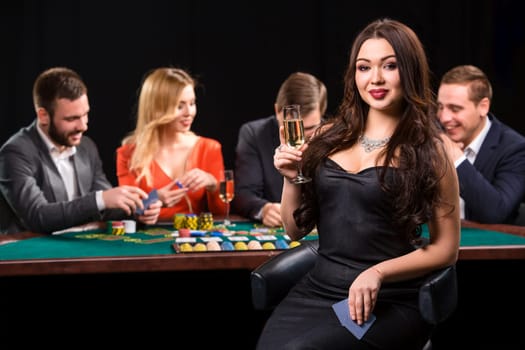 Young people playing poker at the table. Luxury women in dresses with a glass of champagne in hand sitting in the foreground. Casino