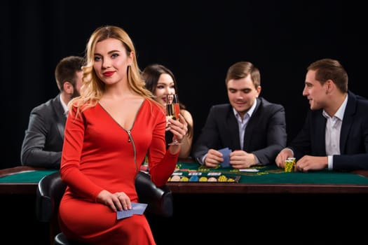 Young people playing poker at the table. Luxury women in dresses with a glass of champagne in hand sitting in the foreground. Casino