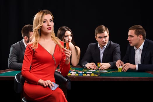 Young people playing poker at the table. Luxury women in dresses with a glass of champagne in hand sitting in the foreground. Casino