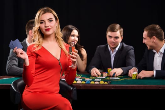 Young people playing poker at the table. Luxury women in dresses with a glass of champagne in hand sitting in the foreground. Casino