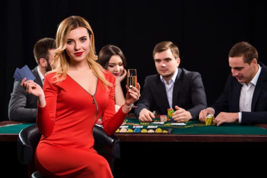 Young people playing poker at the table. Luxury women in dresses with a glass of champagne in hand sitting in the foreground. Casino