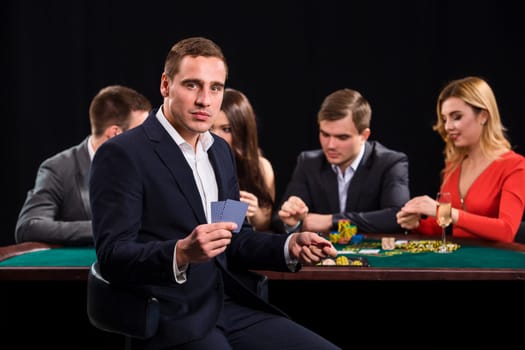 Poker players in casino with cards and chips on black background. A handsome man in the foreground, behind a game table.