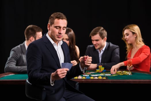 Poker players in casino with cards and chips on black background. A handsome man in the foreground, behind a game table.