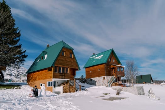 Mom with a small child sit on a sleigh near a snowman in the courtyard of a wooden cottage. High quality photo