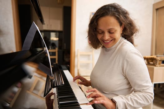 Happy African American woman smiling while playing grand piano, feeling the rhythm of classic music while touching ebony and ivory wooden keys. Female musician practicing the chord musical instrument
