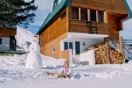 Little girl sits on a sleigh near a snowman next to a wooden cottage. High quality photo