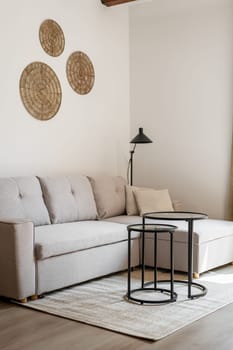 Vertical shot of a beige style corner sofa stands on the floor with a coffee table and circle wall decor and a floor lamp. Concept of a modern living room in a new building.