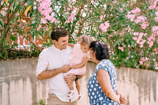 Little girl sits in her father arms, touching her foreheads with her grandmother standing next to her. High quality photo