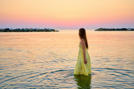 A young woman in a bright yellow dress meets the sunrise, joyfully runs through the sea water