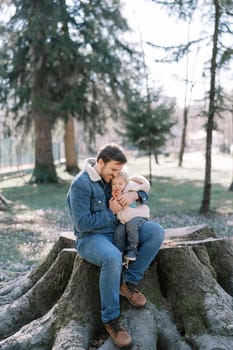 Smiling dad hugging a little girl sitting with her on a big stump in a sunny meadow. High quality photo