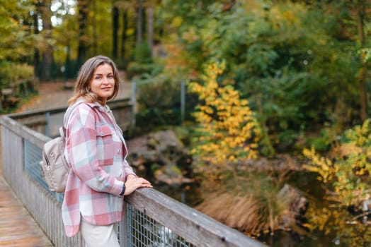 Portrait of cute young woman in casual wear in autumn, standing on bridge against background of an autumn Park and river. Pretty female walking in Park in golden fall. Copy space. smiling girl in the park standing on wooden bridge and looking at the camera in autumn season