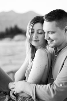 Man hugs woman from behind while sitting with her on the seashore. Black and white photo. High quality photo