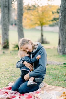 Mom hugs a little boy sitting on her lap on a blanket in the park. High quality photo