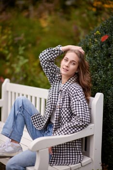 Beautiful teen blonde girl sits on a bench in an autumn park . attractive young smiling girl sitting on a bench in an autumn park. Fall Fashion Serenity: Casual Elegance in the Park
