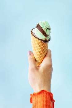 A hand holding an ice cream cone with green and brown scoops against a light blue background