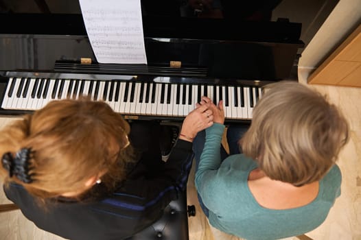 Two mature pretty women musicians engrossed in a music studio lesson, harmonizing melodies on acoustic piano while singing together. People. Lifestyle. Hobbies and leisure activity. View from above