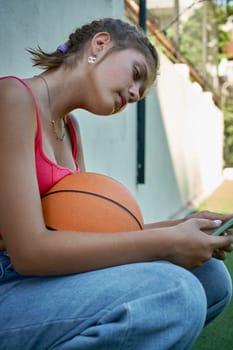 a girl sits with a basketball on the sports ground holding a smartphone in her hands
