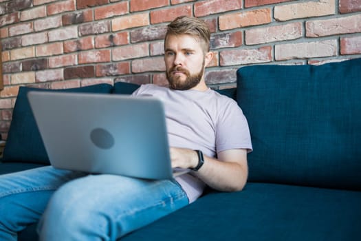Millennial man sitting on sofa at home working on laptop online and using internet. Smiling and happy mood, freelancer and free leisure time, relaxed and modern job lifestyle concept. Copy space and empty place for text.
