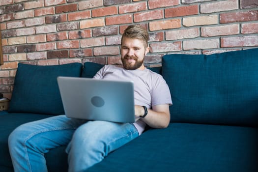 Millennial man sitting on sofa at home working on laptop online and using internet. Smiling and happy mood, freelancer and free leisure time, relaxed and modern job lifestyle concept. Copy space and empty place for text.