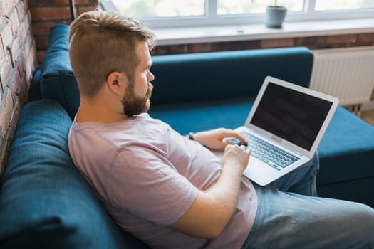 Millennial man sitting on sofa at home working on laptop online and using internet. Smiling and happy mood, freelancer and free leisure time, relaxed and modern job lifestyle concept. Copy space and empty place for text.