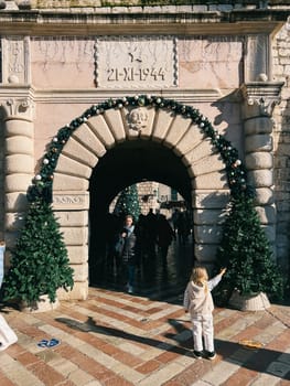 Little girl stands in front of an ancient arch and points to a decorated Christmas tree. High quality photo