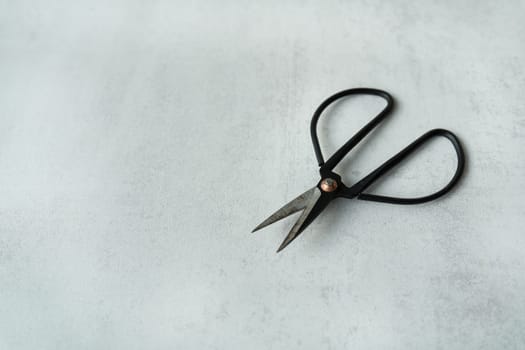 A black scissors with pointed tips lying on a textured grey surface. The handles of the scissors are round and appear to be made of metal