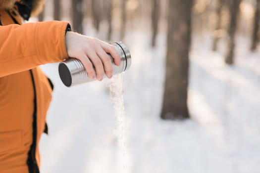 Man pours hot water from a thermos into a snow walking in snowy frozen winter forest at sunset. Adventure, tourism and camping concept. Copy space and empty place for text advertising.