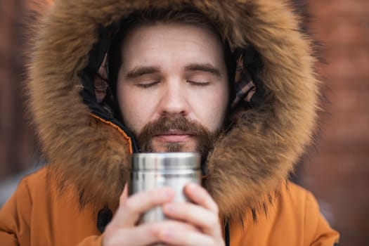 Close-up portrait handsome bearded millennial man in winter clothes and with thermos snow outdoor. Cold season and hot beverage in winter time.