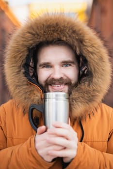 Close-up portrait handsome bearded millennial man in winter clothes and with thermos snow outdoor. Cold season and hot beverage in winter time.