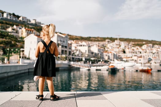 Little girl stands on the pier and looks at the colorful villas with moored yachts. Back view. High quality photo