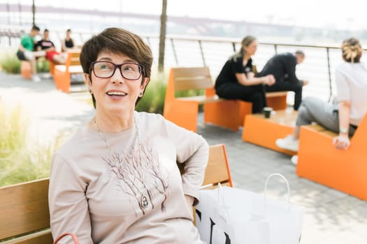 Outdoor portrait of beautiful and elegant middle age 55 - 60 year old woman wearing long sleeve top sitting on bench on the street. Copy space and empty place for text advertising.