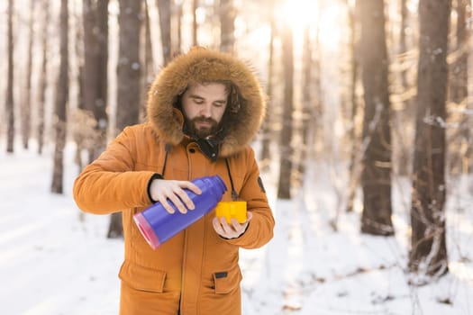 Man pours hot tea from a thermos into a snow walking in snowy frozen winter forest at sunset. Adventure, tourism and camping concept. Copy space and empty place for text advertising.