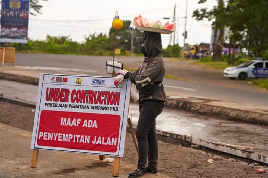 A woman with a tray full of food on her head sells snacks along the road near a road repair sign on a major highway in Asia. Bali, Indonesia - 12.11.2022