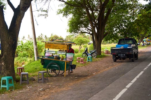 A street food vendor stands working on the side of the road in an Indonesian village in Bali. Bali, Indonesia - 12.11.2022