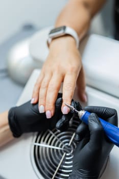 Female master using electric nail file while doing manicure for client in beauty salon.