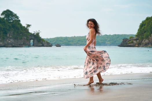 A slender young woman in a pink light dress walks along a tropical beach. The girl is dancing in the sea waves