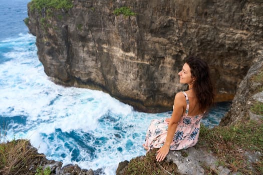 A beautiful woman in a pink dress sits on a cliff above the ocean on the island of Nusa Penida. Devil's Billabong an incredibly wonderful lagoon with splashes from the waves