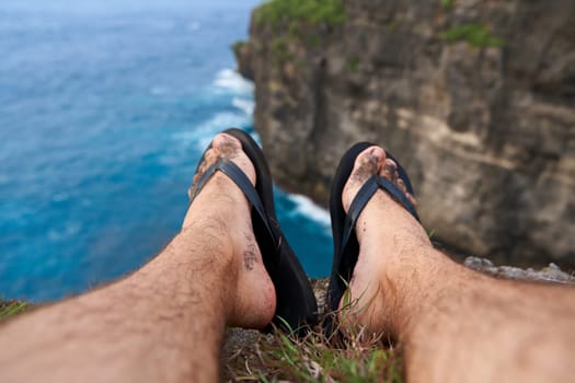 A man sits on a cliff with his legs dangling over an abyss above the ocean. First person photo
