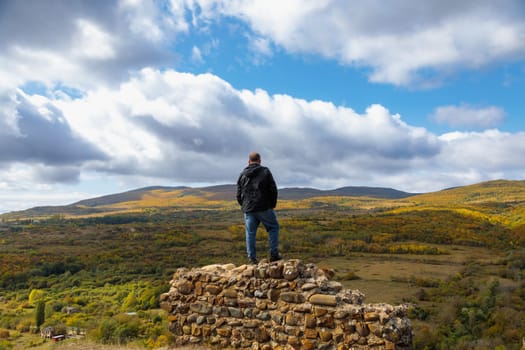 A man is finding peace as he stands on a ruined fortress wall, admiring the breathtaking and serene mountain scenery that lies before him on a beautiful day.