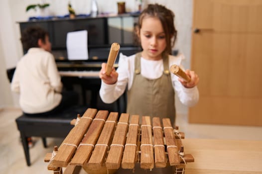Little Caucasian child girl beating with wooden mallets on xylophone, playing music against her brother sitting at grand piano. Kids education and entertainment. Music lesson. Leisure and hobbies