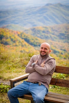 This captivating image captures a serene moment as a man relaxes on a rustic bench, taking in the grandeur of the mountainous terrain with its jagged peaks and tranquil atmosphere.